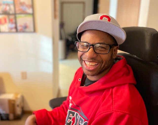 Tyrone, wearing Ohio State sweatshirt and hat, sitting in wheelchair smiling