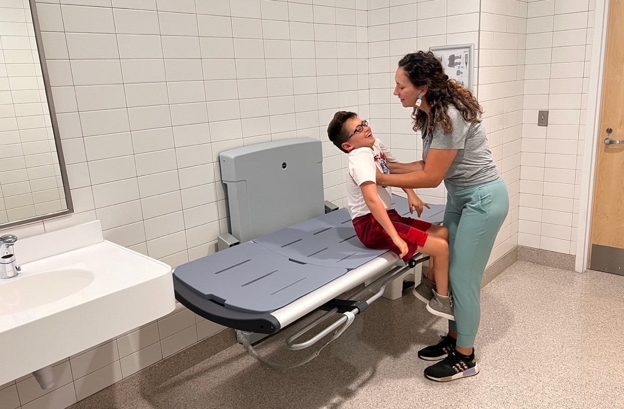 A mother is lifting her 9-year-old son onto a large changing table in a hospital restroom.