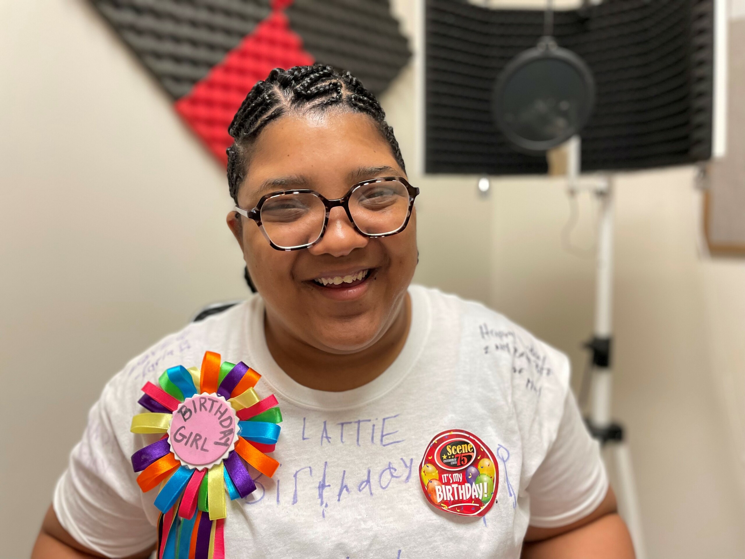 Smiling woman in white t-shirt with colorful birthday girl ribbon. She is sitting in a recording studio.