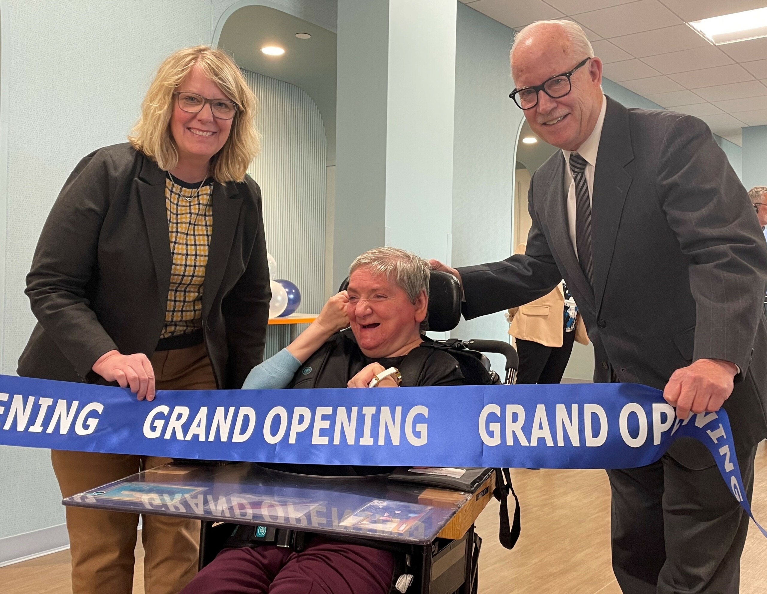 State DODD director Kim Hauck holding blue grand opening ribbon next to woman in wheelchair and county disabilities board Superintendent Jed Morison. Morison holding other end of ribbon.